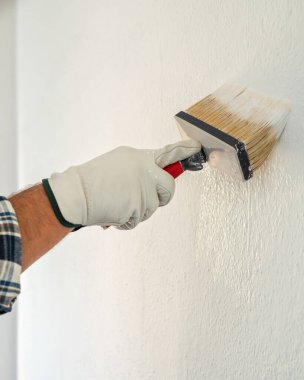 Koruyucu eldiven takan ellerin yakın çekimi. Bir ressam boya fırçasını kullanarak duvarları beyaz boyayla boyar. İnşaat endüstrisi. İş Güvenliği.