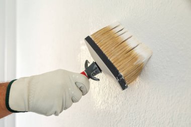 Koruyucu eldiven takan ellerin yakın çekimi. Bir ressam boya fırçasını kullanarak duvarları beyaz boyayla boyar. İnşaat endüstrisi. İş Güvenliği.