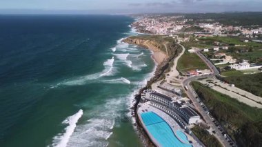 Portekiz 'deki Praia Grande sahil şeridine ait 4K hava görüntüleri, okyanus dalgalarını, büyük bir havuzu ve güneşli bir günde kasabayı çevreleyen uçurum kenarındaki oteli gösteriyor..