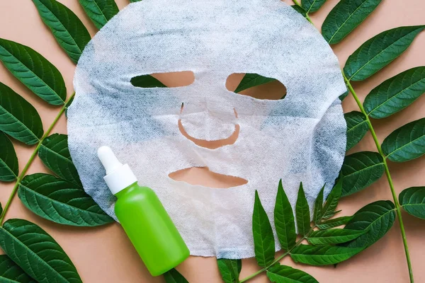 Fabric cosmetic face mask and serum bottle on green leaves. Spa beauty treatments for facial skin care. Flat lay.