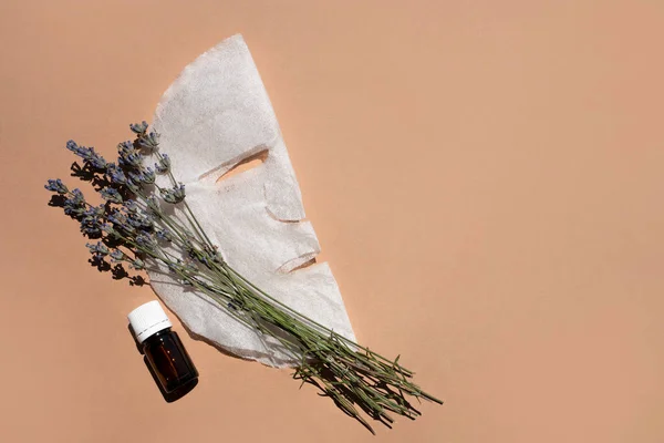 stock image Fabric face mask, organic oil vial and lavender bouquet on beige background. Flat styling, space for text.
