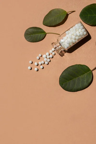 Homeopathic glass bottle with pills and natural leaves on a beige background. Alternative medicine of medicinal herbs, homeopathy. Vertical image, place for text.