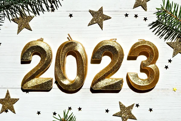 Holiday background Happy New Year 2023. Golden sparkling numbers 2023 on a white wooden background with fir branches. Side view, close-up.