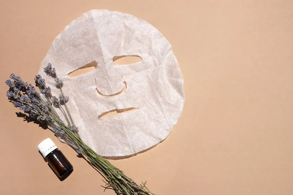 Fabric face mask, organic oil vial and lavender bouquet on beige background. Flat styling, space for text.