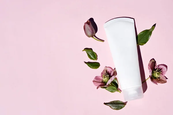 stock image Natural cosmetic cream in white tube, skin care mockup concept on pink background with natural hellebore flowers. a flat lay, place for text.