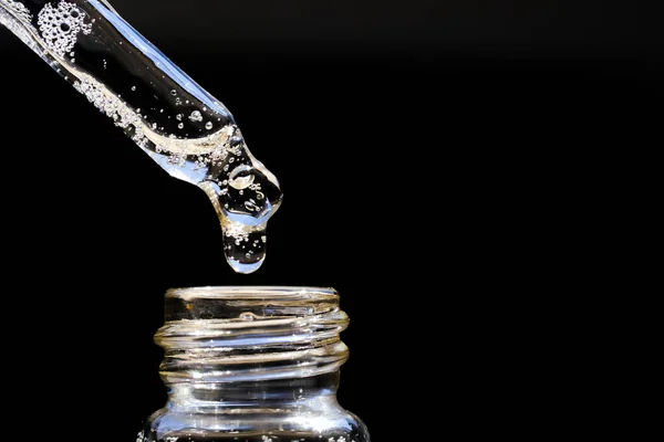 A drop of serum with air bubbles drips from a pipette into a glass bottle on a black background. Front view and close-up.