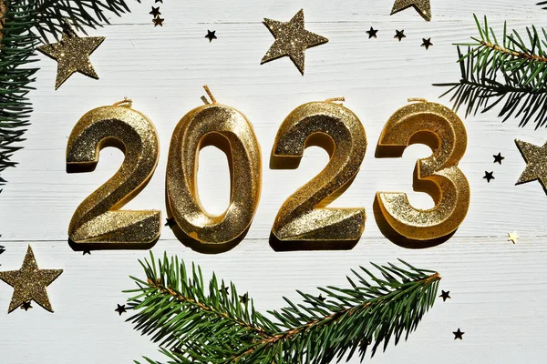 Holiday background Happy New Year 2023. Golden sparkling numbers 2023 on a white wooden background with fir branches. Side view, close-up.