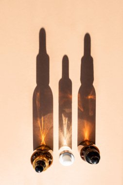 Bej arka planda arka arkaya dizilmiş üç cam şişe kozmetik sıvısı, gölgeler. Dikey resim, üst görünüm.