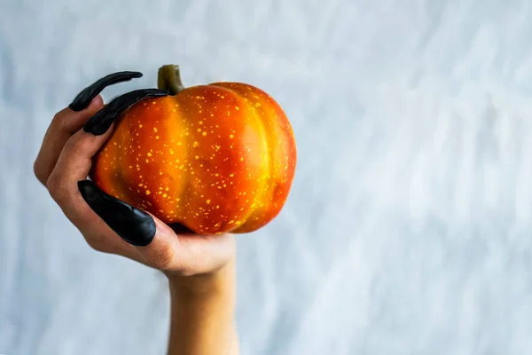 stock image Halloween theme. A hand with black long nails holds a pumpkin against the background of a white sheet. Side view, place for text.