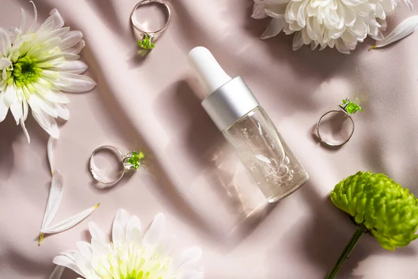 A transparent bottle with a natural cosmetic product on a pastel pink background with natural flowers. Skin care concept. Flat lay.