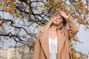 Uzun beyaz saçlı güzel bir kadın, sarı yapraklı, bej ceketli bir ağacın yanındaki parkta duruyor..