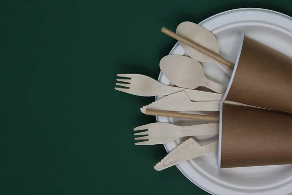 Disposable bamboo cutlery in disposable paper cups on dark green background. Zero waste concept.