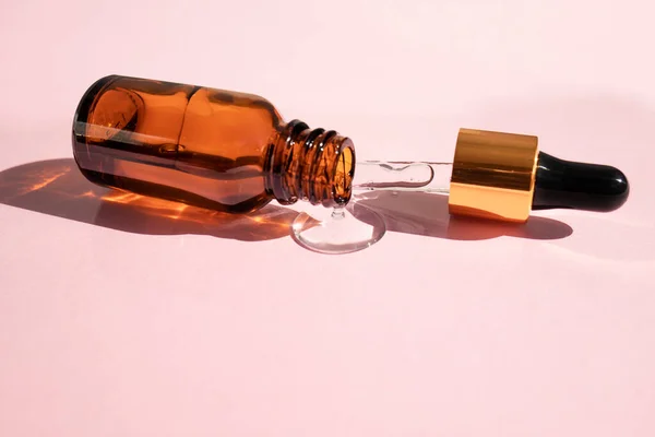 Glass dropper bottle with dropper and spilled cosmetic liquid on pink background. Side view, space for text.