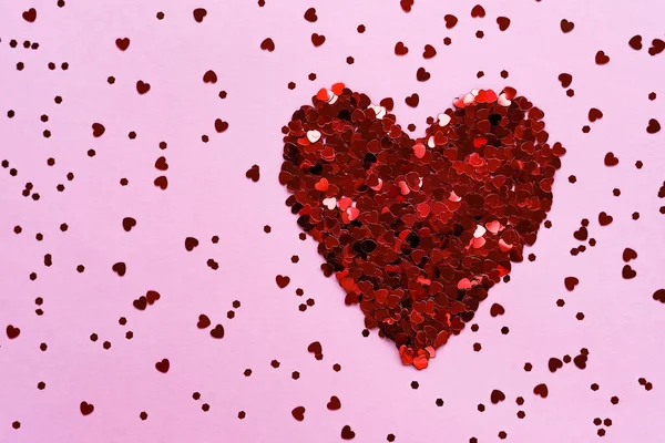 Heart made of shiny red small decorative hearts on a pink background strewn with sparkles. Flat lay, place for text. Concept for valentine's day.