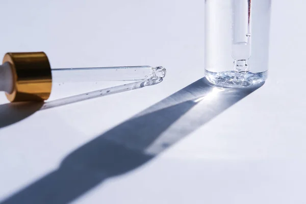 A glass pipette and a transparent bottle of cosmetic liquid cast a shadow on a white background. Side view.