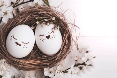 Yuvalarında şirin yüzleri olan Paskalya yumurtaları ve beyaz ahşap arka planda doğal kiraz çiçekleri dalları. Paskalya konsepti. Üst görünüm, metin için boşluk.