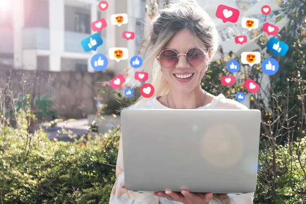 A happy blonde with a laptop gets emoji and emoticons for her online post. The concept of social media.