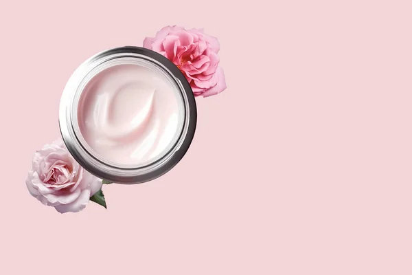 Facial cream in a glass jar with powdery rose flowers on a light pink background. Cosmetics with hyaluronic acid. Natural branding skin care product. Flat lay, top view, copy space.