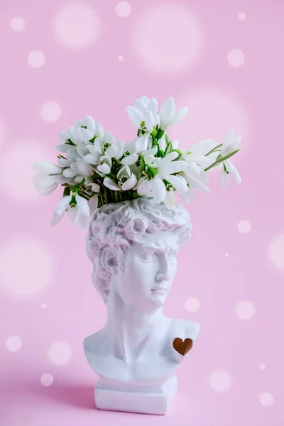 White plaster statue of David\'s head with snowdrops on pastel pink background. Vertical image.