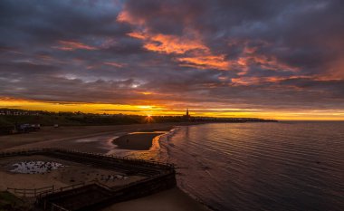 Dramatik bir günbatımı Tynemouth Longsand plajında bulutlu gökyüzünü aydınlatıyor İngiltere 'nin kuzeydoğusundaki CullerCoats' a doğru bakıyor.