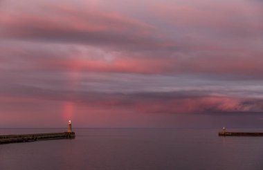 A dramatic sunset lights up the cloudy sky as a vibrant rainbow appears in Tynemouth in the North East of England clipart