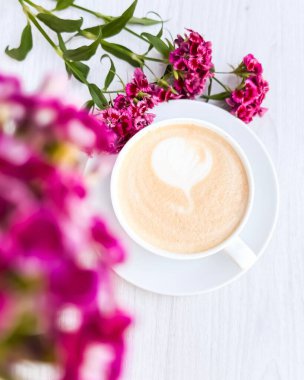 Beyaz Tahta Masa Üzerinde Kalp Şeklinde Köpük ve Taze Çiçeklerle Bir fincan Latte.