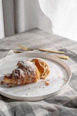 Kareli keten peçetenin üzerinde beyaz bir tabakta servis edilen pudra şekerli taze pişmiş kruvasan. Yumuşak gölgeli doğal sabah ışığı altında yakalanmış, rahat ve minimal bir kahvaltı sahnesi yaratıyor. Yemek tasarımı ve fırın için ideal.