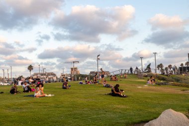 Gün batımında kumsalın yanındaki parkta dinlenen, kitap okuyan, gün batımını izleyen, konuşan, yoga yapan insanlar.