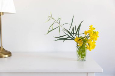 Tecoma 'lı çalı dalı, iç taraftaki beyaz bir masada cam bir şişede çiçek açar..