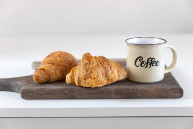 İki kruvasan, üzerinde kahve yazılı bir fincan beyaz bir masanın tahta tahtasına. Metin için yer