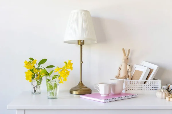 White floor lamp with lampshade. A pink notebook with springs, a white coffee mug, a wooden toy bunny on hinges, sitting on a plastic box. Dresser. Minimalism. Seventies style.