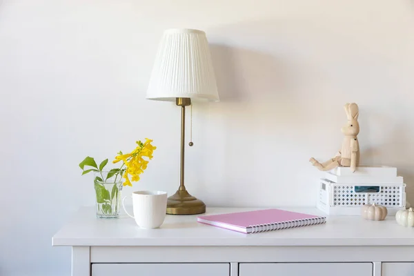 White floor lamp with lampshade. A pink notebook with springs, a white coffee mug, a wooden toy bunny on hinges, sitting on a plastic box. Dresser. Minimalism. Seventies style.