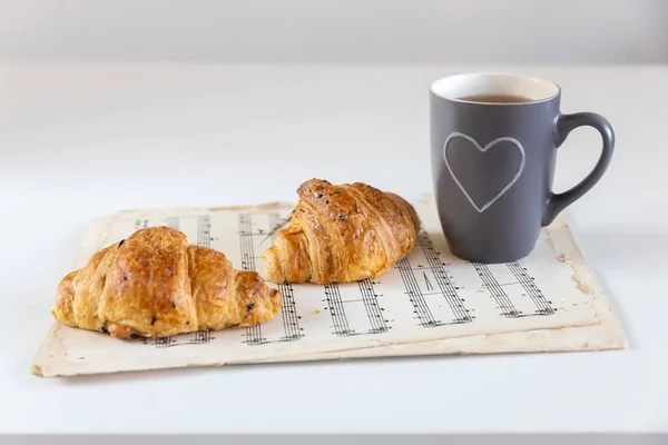 Two croissants with chocolate chips on a white sheet of paper on the table, a gray cup with a white heart with tea. Place for text