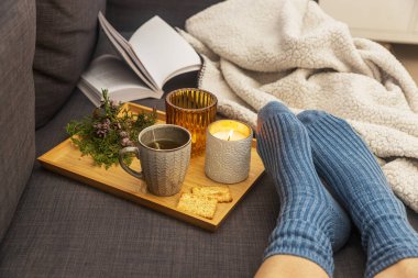 Kanepenin üzerinde yün mavi çoraplar içinde bir kadın bacakları, bir kitap, bir fincan çay ve tepside bir mum. İç ve ev konforu. Üst görünüm
