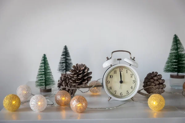 A white alarm clock, a twelve pointer, a small artificial green Christmas tree and garland on a white chest of drawers. Copy space. Christmas card.