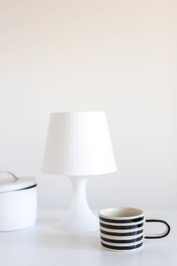 A black and white striped mug, a lamp with a shade and a cookie box on a white table. Vertical frame. Space for text.