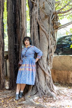 Gülümseyen esmer kız, yirmi beş yaşında klasik mavi desenli bir elbise giyip parktaki büyük bir incirin gövdesine yaslanmış küçük bir çanta taşıyor.