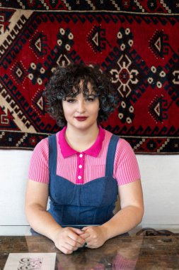 25 yaşında, esmer, gülümseyen bir kızın portresi. Pembe bir bluz ve mavi kadife bir tulum giymiş. Bir kafedeki kırmızı Türk halısının yanındaki cam bir masada oturuyor..