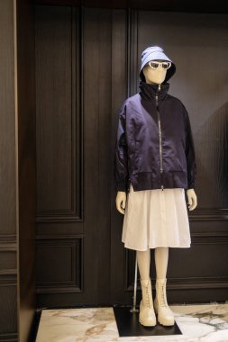 Manken koyu mavi bir pilot ceketi, beyaz bir etek, panama şapkası, güneş gözlüğü ve siyah bir duvara karşı beyaz bir spor ayakkabı giyiyor..