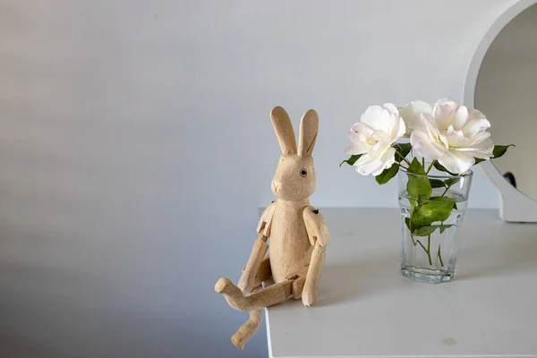 Tea white rose in a transparent glass on a white table. There is wooden hare on hinges. Place for text.