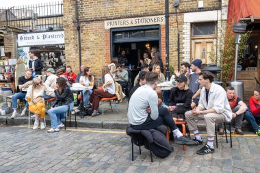 Londra, İngiltere - 17 Nisan 2023: Columbia Road Çiçek Pazarı. Yerel halk iyi havada kaldırımda otururken kahve ve alkol içer.