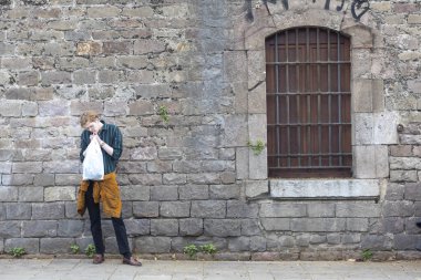 Evdeki tuğla duvarın yanında sigara yakan genç bir adam.