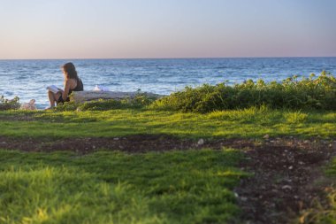 Hayfa, İsrail - 27 Eylül 2023, Uzun saçlı genç bir kız Bat Galim sahilinde gün batımında çimlerin üzerinde oturan bir kitap okuyor..