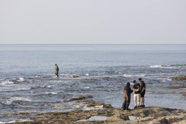 Hayfa, İsrail - 11 Ocak 2024, gezinti güvertesi boyunca deniz boyunca yürü. İnsanlar sette oturur, denize bakar, arkadaşlarıyla sohbet ederler. Balıkçılar sığ sularda balık yakalar