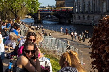 Berlin, Almanya - 02 Nisan 2024, İnsanlar setin üzerinde oturuyorlar. Berlin 'de ilkbahar. Neşeli insanlarla dolu bir eğlence gemisi kanal boyunca yol alıyor..