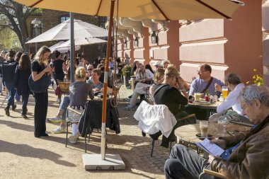 Berlin, Almanya - 20 Nisan 2024 İnsanlar açık bir kafede öğle yemeği yiyor ve nehir kenarındaki bir parkta bahar güneşinin tadını çıkarıyorlar..