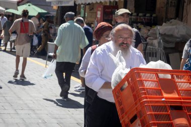Kudüs, İsrail - 1 Ekim 2024, Kippa ve beyaz gömlekli yaşlı bir adam, Ortodoks bir Yahudi, Mahane Yehuda pazarındaki kalabalığın arasından kırmızı meyve kutularını önüne atıyor..