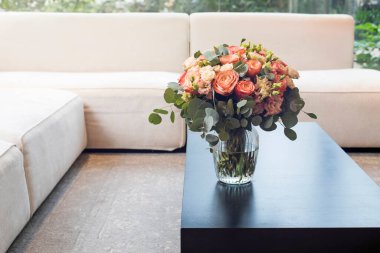 Bir buket turuncu gül, pembe ve yeşil lisianthus, ve okaliptüs dalları pencereye bakan beyaz bir kanepenin yanındaki sehpanın üzerinde duruyor. Minimalizm. İç dekorasyon.