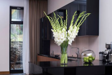 A white bouquet of gladioluses on the kitchen table in a tall glass vase with a bowl of green apples. clipart