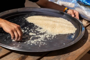 Bir adam, tahta bir masada pilav yaparken büyük çelik bir tepside pirinci sınıflandırır..